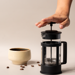 Hand pressing down the plunger of a French press coffee maker beside a ceramic cup and scattered coffee beans.