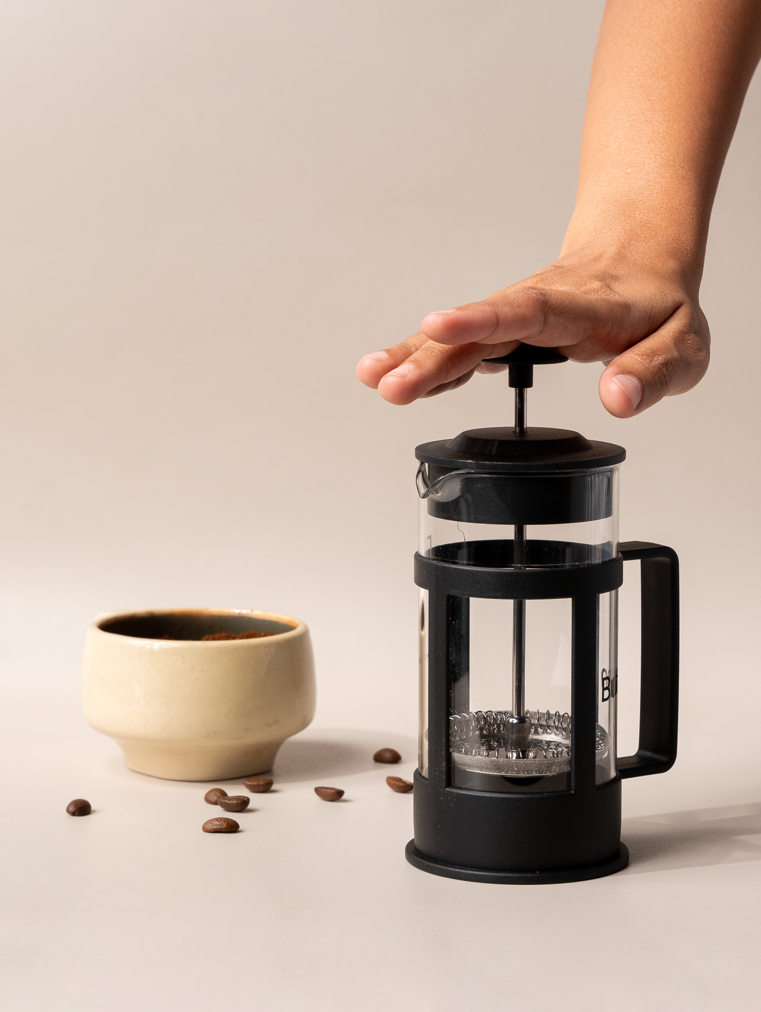 Hand pressing down the plunger of a French press coffee maker beside a ceramic cup and scattered coffee beans.