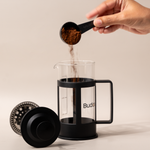 Hand pouring ground coffee into a Budapresso French press, ready for brewing fresh specialty coffee.