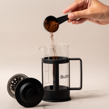 Hand pouring ground coffee into a Budapresso French press, ready for brewing fresh specialty coffee.