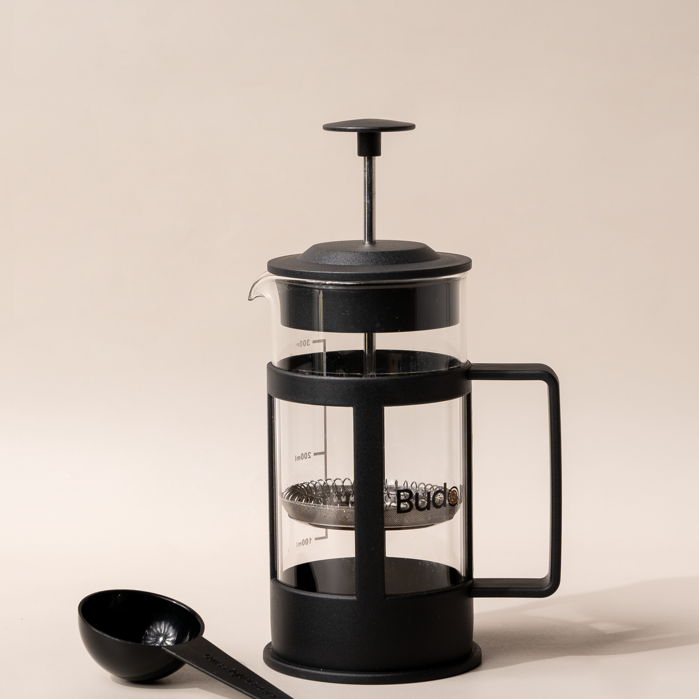 A black French press coffee maker with a glass body and a metal scoop on a light background.