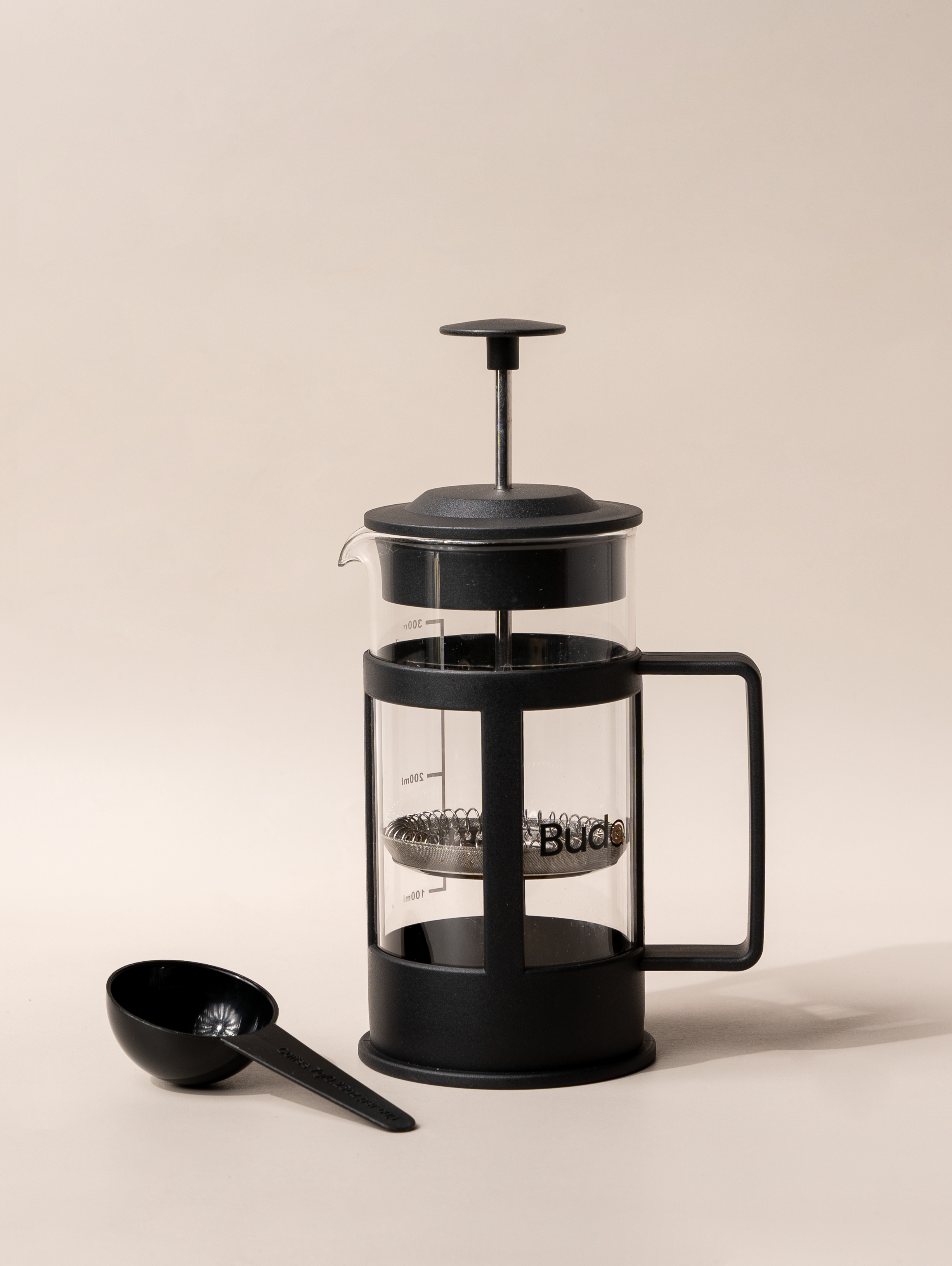 A black French press coffee maker with a glass body and a metal scoop on a light background.
