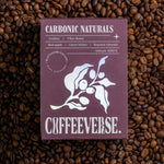 A package of Carbonic Naturals coffee with a purple label, displaying the brand name and coffee variety, placed on a background of coffee beans.